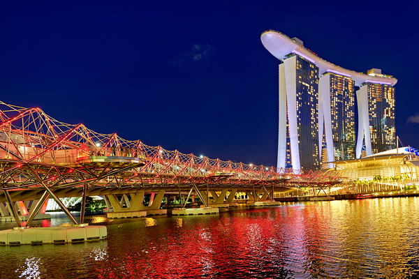 People walk across the Helix Bridge as the Marina Bay Sands Hotel shines at sunset. The sky turns orange and lights reflect in the water. Digital Download