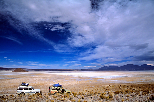 Vehicles traverse a vast arid salt flat landscape. Digital Download