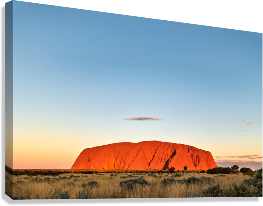 Uluru glows orange at sunset under a clear blue sky.