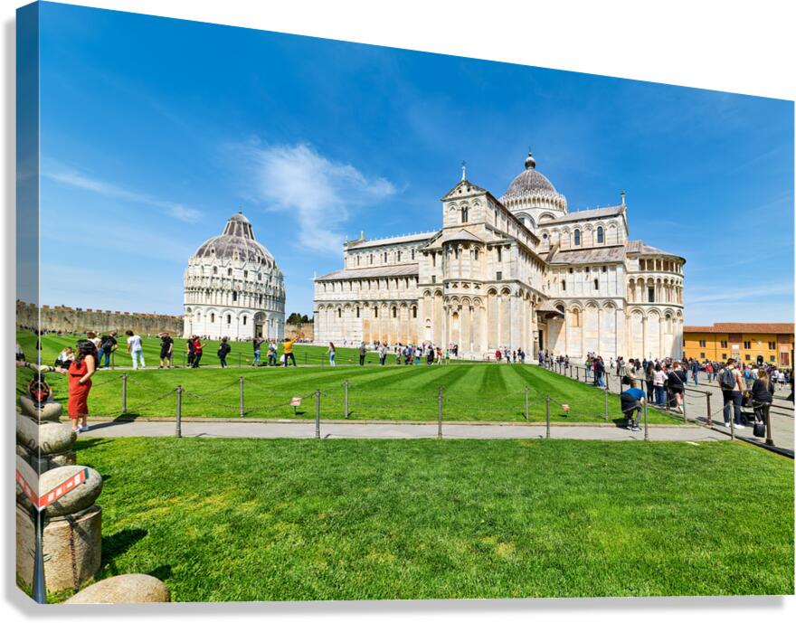 People walk in Piazza dei Miracoli admiring the Baptistry and Cathedral in Pisa. The sun shines on the historic buildings and green grass.