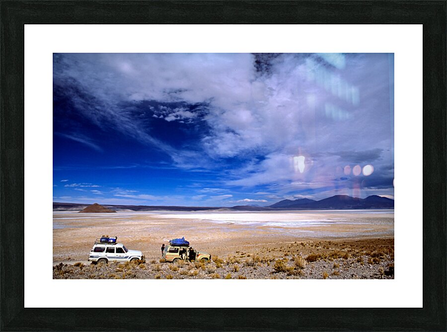 Vehicles traverse a vast arid salt flat landscape. Picture Frame print