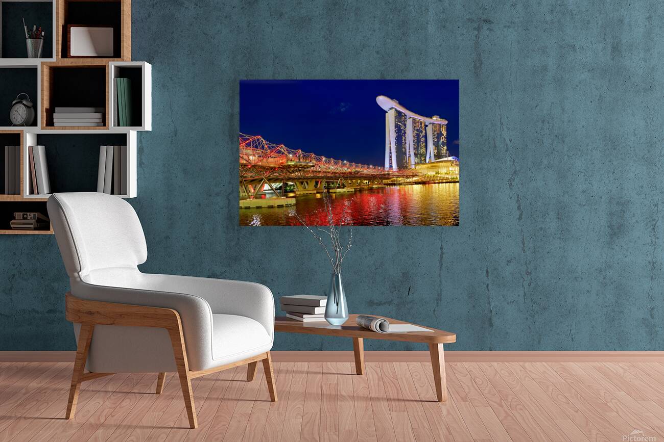 People walk across the Helix Bridge as the Marina Bay Sands Hotel shines at sunset. The sky turns orange and lights reflect in the water. Reproduction