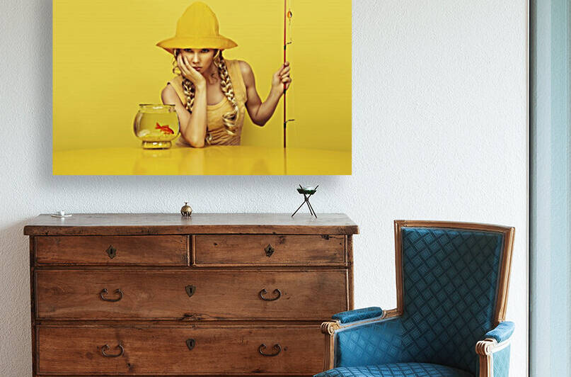 Portrait of a young bored fisherwoman her chin resting on her hand dressed in a yellow dress with a fish pattern fishing a goldfish in its fishbowl placed on a yellow table by Artmood Visualz