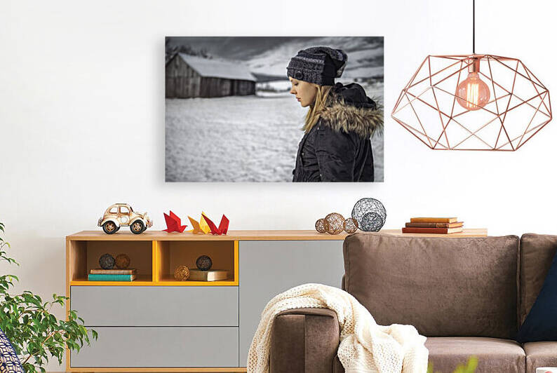 Side view of a beautiful blonde woman looking down wearing a black winter coat and a Beanie hat standing alone in a snowy winter rural landscape with a cloudy sky by Artmood Visualz