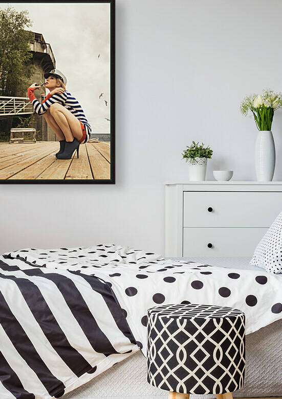 Attractive young blonde woman wearing a striped romper and a captains hat smoking a pipe while squating on the ground of a harbour by Artmood Visualz