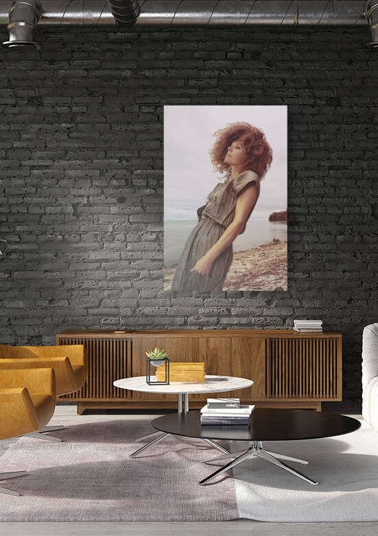 Beautiful young woman with brown curly hair on a pebble beach by the water on a windy and cloudy day by Artmood Visualz
