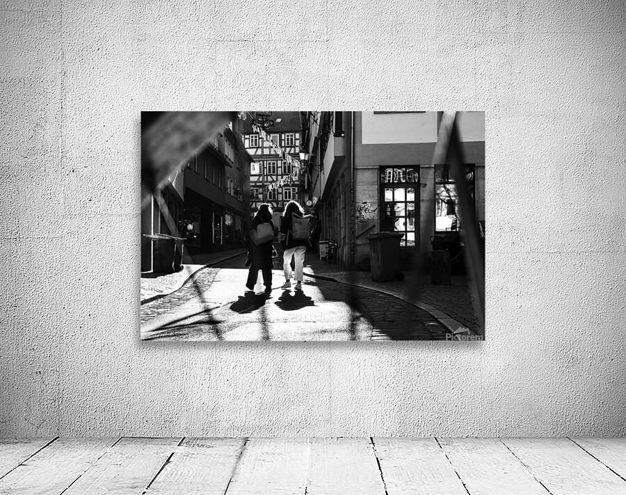 Black and white street photo of people walking down the road seen through the spokes of a bicycle Wall Preview